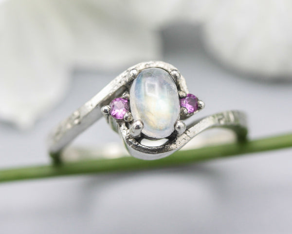 Oval cabochon moonstone ring with pink sapphire side set gems in prongs setting with sterling silver oxidized texture band