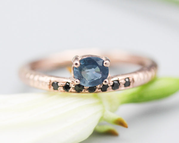 Oval faceted blue sapphire ring in prongs setting with tiny black spinel on 14k Rose gold texture design band