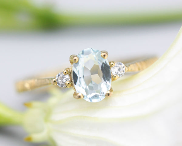 Oval faceted blue topaz ring with tiny round diamonds side set gems in prongs setting with 14k gold texture band